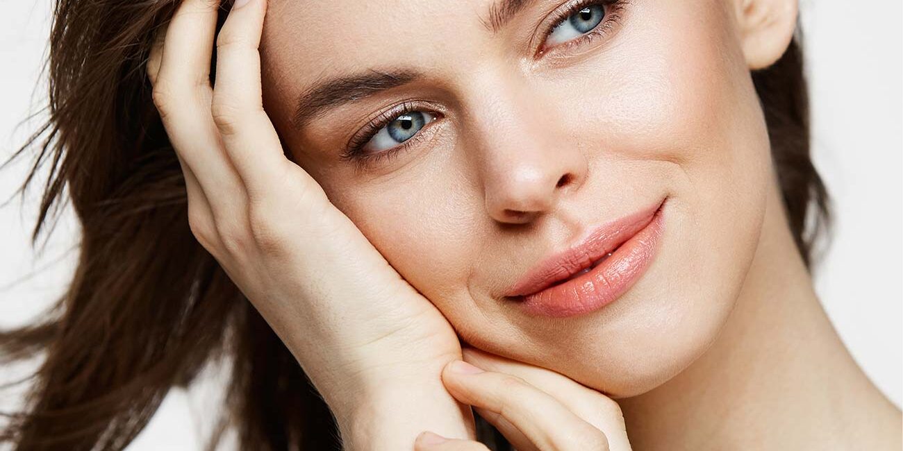 Close-up of a smiling young woman with clear skin, gentle expression, and brown hair touching her face.