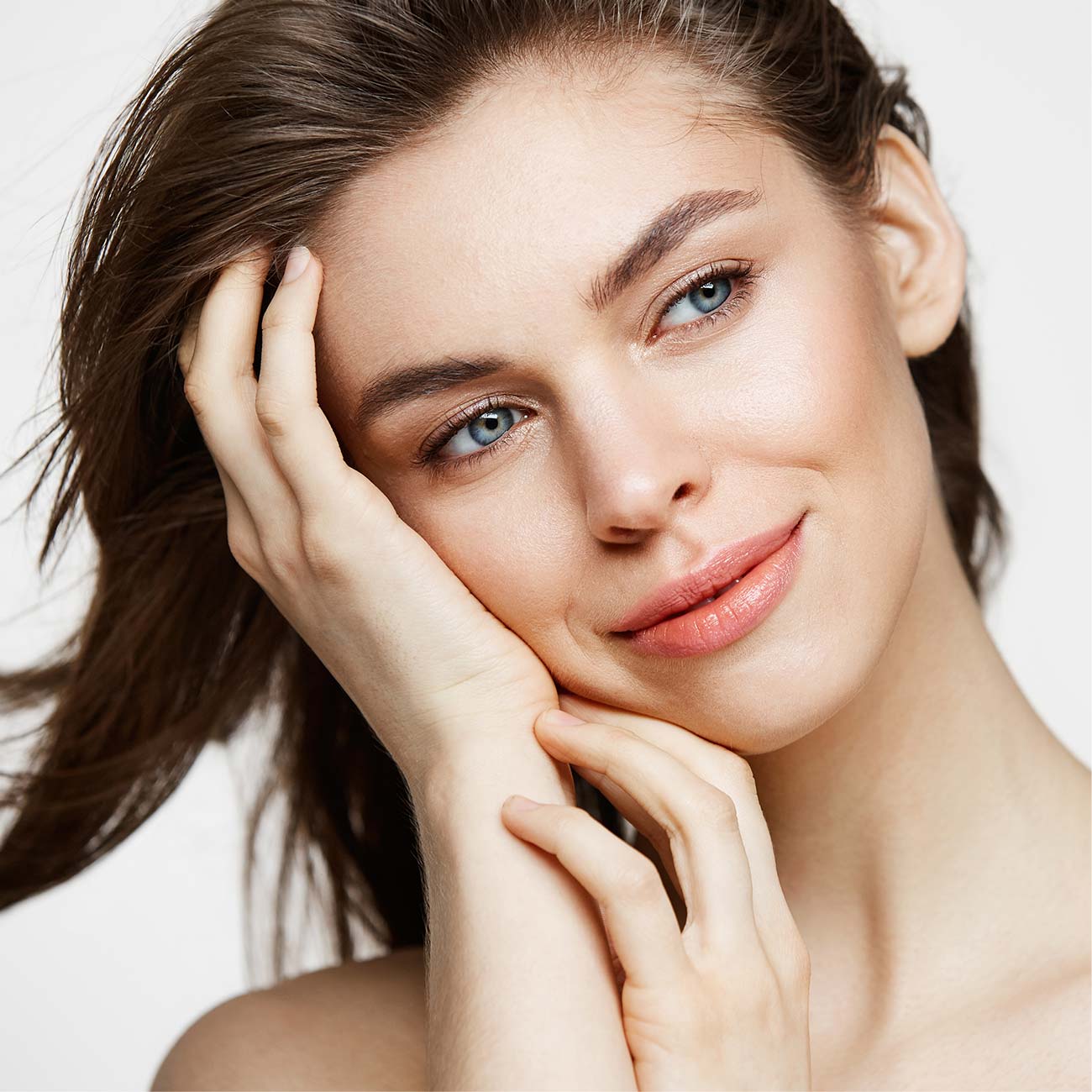 Close-up of a smiling young woman with clear skin, gentle expression, and brown hair touching her face.